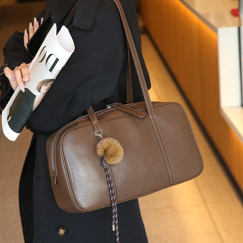 Brown Teak worn on the shoulder over a black coat, with a pom-pom charm hanging from the zip